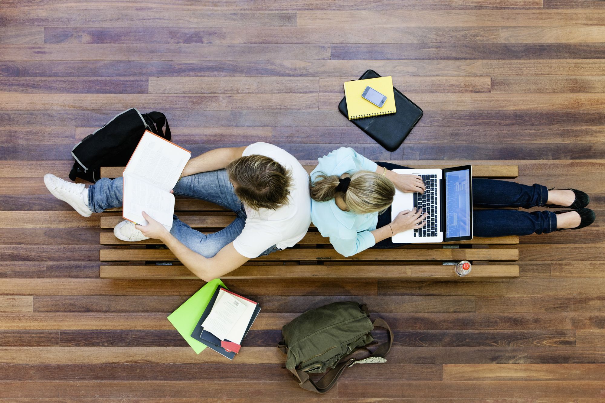 Two students sit back to back on a bench, studying, reading and typing on a laptop.