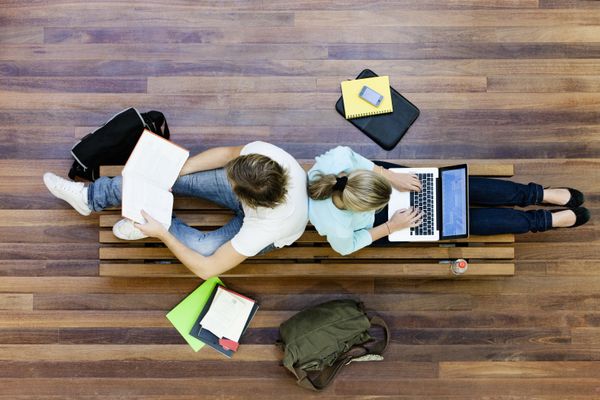 Two college students sit back to back on a bench while he reads and she types on a laptop.